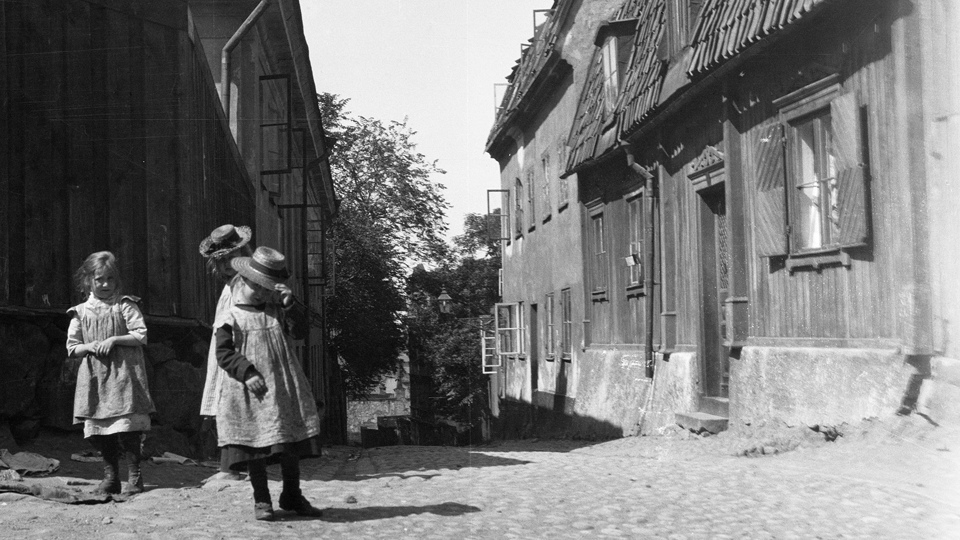 Flickor i gammeldags kläder står på en kullerstensgata bland trähus.